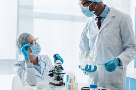 multicultural biologists in white coats and medical masks talking in lab