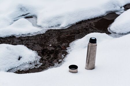 Vacuum flask on snow near flowing mountain streamの写真素材