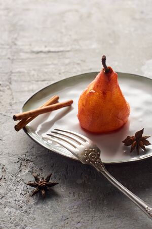 delicious pear in wine with cinnamon and anise on plate with silver fork on grey concrete surfaceの写真素材