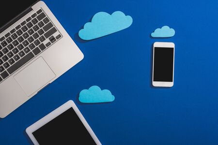 top view of empty paper clouds near laptop, smartphone and digital tablet with blank screen isolated on blueの写真素材