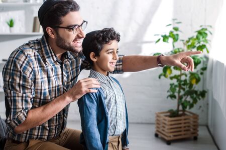 smiling jewish father pointing with finger and looking through window with son in apartmentの写真素材