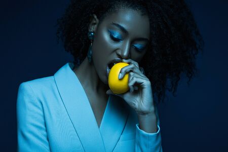 attractive african american woman with makeup eating lemon isolated on blueの写真素材