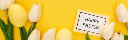 top view of tulips and painted Easter eggs near card with happy Easter lettering on colorful yellow...