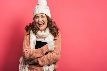 cheerful girl in warm hat and scarf holding digital tablet on pink backgroundの写真素材