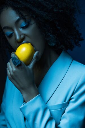 attractive african american woman with makeup eating lemon isolated on blueの写真素材
