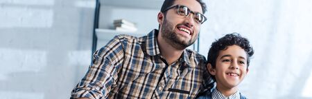 panoramic shot of smiling jewish father and son watching tv in apartment