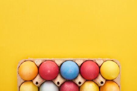 top view of multicolored painted Easter eggs in cardboard container on yellow background with copy spaceの写真素材