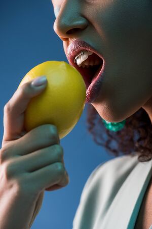 cropped view of african american woman with braces eating lemon isolated on blueの写真素材