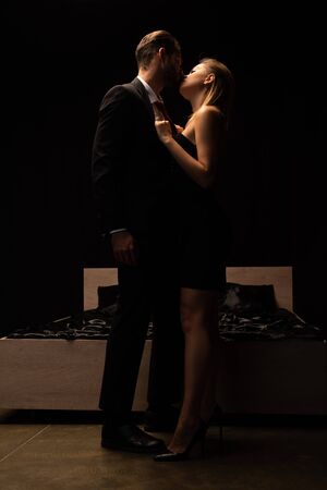 beautiful passionate couple kissing and standing near bed in dark roomの写真素材