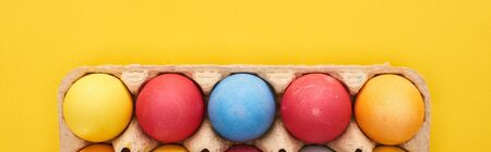 top view of multicolored painted Easter eggs in cardboard container on yellow background, panoramic...