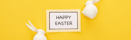 top view of greeting card with happy Easter lettering near Easter white bunnies on yellow colorful background, panoramic shotの写真素材