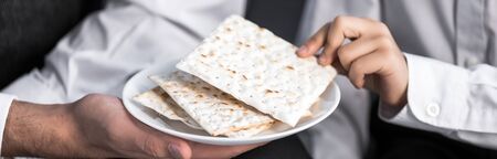 panoramic shot of jewish father holding plate and son taking matza in apartment 