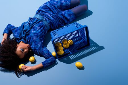 high angle view of african american woman lying with shopping basket and lemons on blue backgroundの写真素材