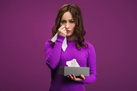 sad woman wiping tears with paper napkin while crying on purple background