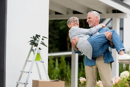 mature man holding smiling woman in glasses near new houseの写真素材