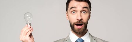 panoramic shot of excited businessman holding light bulb and looking at camera isolated on greyの写真素材