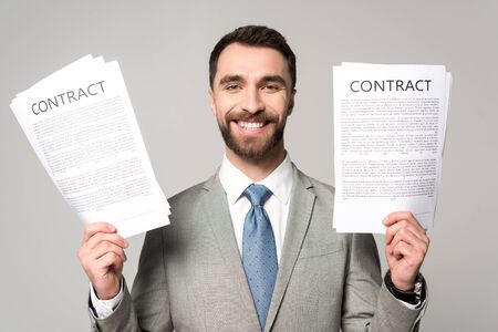 happy businessman holding contracts and looking at camera isolated on greyの写真素材