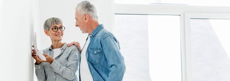 panoramic shot of smiling man looking at mature woman with palette in new house