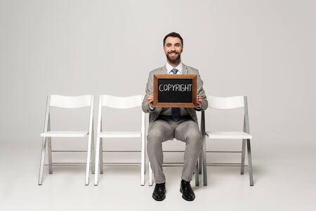 happy businessman holding chalkboard with word copyright while sitting on chair on grey background