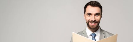 panoramic shot of positive businessman smiling at camera while holding paper folder isolated on greyの写真素材