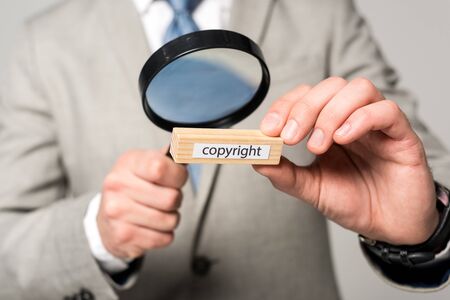 cropped view of businessman looking at wooden block with word copyright through magnifier isolated on greyの写真素材