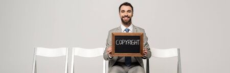 panoramic shot of happy businessman holding chalkboard with word copyright while sitting on chair isolated on greyの写真素材
