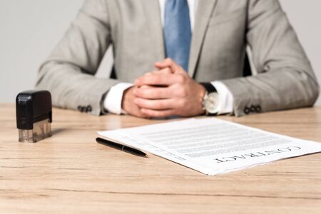cropped view of businessman sitting at desk near contract, stamp and pen isolated on grey