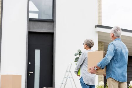 mature man holding box and woman holding plant near new houseの写真素材