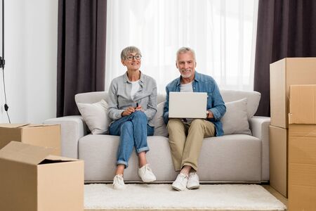 mature man with laptop and woman smiling at camera in new houseの写真素材