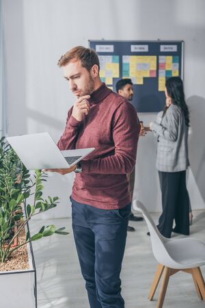 selective focus of handsome scrum master using laptop near multicultural coworkers