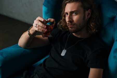 fashionable businessman in black outfit holding glass of whiskey in armchair