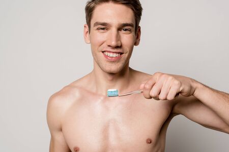 smiling sexy man with bare torso holding toothbrush isolated on grey