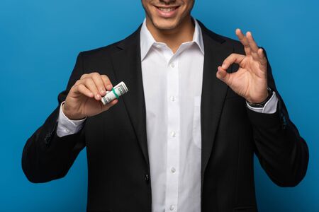 Cropped view of smiling businessman showing okay gesture and holding cash roll on blue backgroundの写真素材