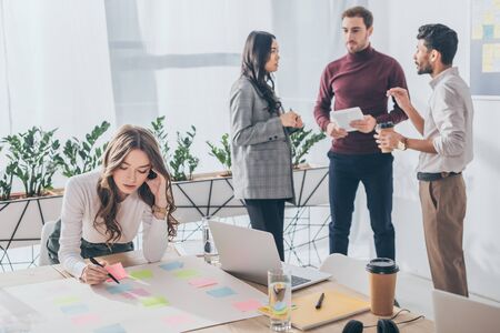 selective focus of attractive businesswoman writing on sticky note near multicultural coworkers