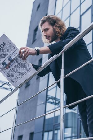 low angle view of fashionable businessman in black suit reading business newspaper near office building and railingの写真素材