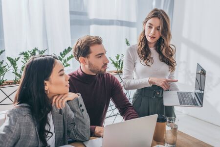 attractive businesswoman pointing with finger at laptop near multicultural coworkers
