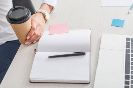 cropped view of businesswoman holding paper cup near notebook and laptop in officeの写真素材