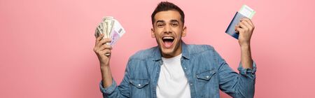 Panoramic shot of excited man holding cash, passport and boarding pass on pink background