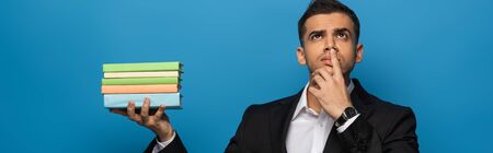 Panoramic shot of pensive businessman holding books and looking away isolated on blueの写真素材
