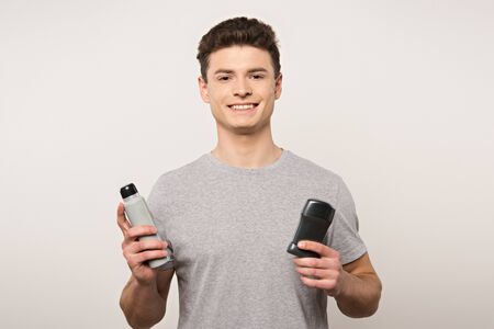 young man in grey t-shirt holding deodorants and smiling at camera isolated on greyの写真素材
