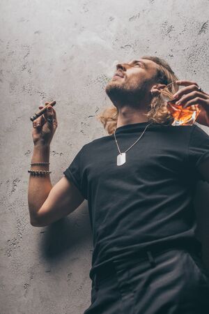 low angle view of fashionable businessman in black outfit smoking cigar and drinking whiskey near...