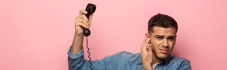 Panoramic shot of confused man holding telephone handset and closing ear isolated on pink