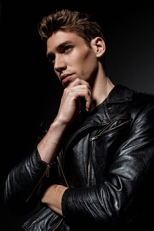 sexy young man in biker jacket touching chin and looking away on black backgroundの写真素材