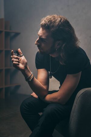 fashionable businessman in black outfit sitting in armchair with lighter near grey wall