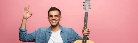 Panoramic shot of man with acoustic guitar smiling at camera and showing okay isolated on pink