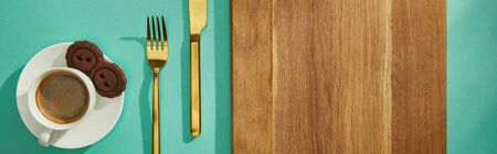 Top view of coffee cup with cookies on saucer, golden cutlery and cutting board on turquoise surface, panoramic shotの写真素材