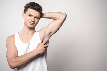 handsome man in white sleeveless shirt applying deodorant on underarm and smiling at camera on grey backgroundの写真素材