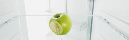 Panoramic shot of bitten off green apple on shelf of refrigerator with open door isolated on whiteの写真素材