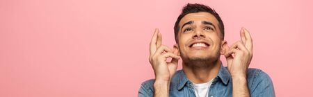 Panoramic shot of smiling man with crossed fingers looking away isolated on pink