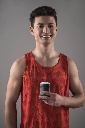 young man in red sleeveless shirt holding deodorant while smiling at camera isolated on greyの写真素材
